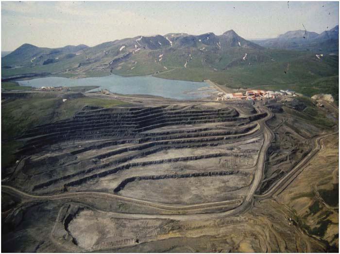 A large section of land has been removed by strip mining near a lake with mountains in the background 