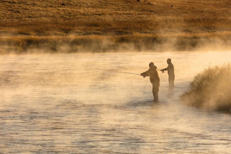 Two anglers fishing on a river at sunrise, with mist rising from the river around them