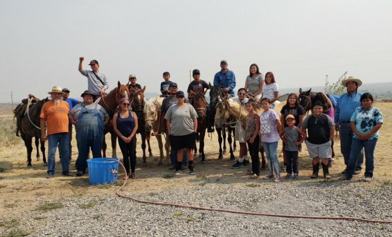 A group of indigenous people, some sitting on horses and some standing in front, in front of a prairie